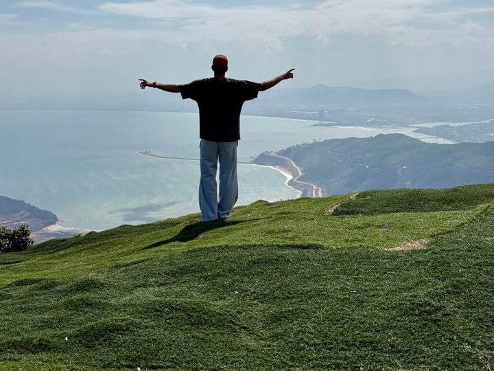 Pablo seen from behind with arms outstretched on a green hillside facing the sea
